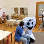 Ein Lehrling der zahnärzlichen Fachassistenz zeigt anhand eines als Zahn verkleideten Mitschülers wie die richtige Zahnpflege mit einer übergroßen Zahnbürste funktioniert.